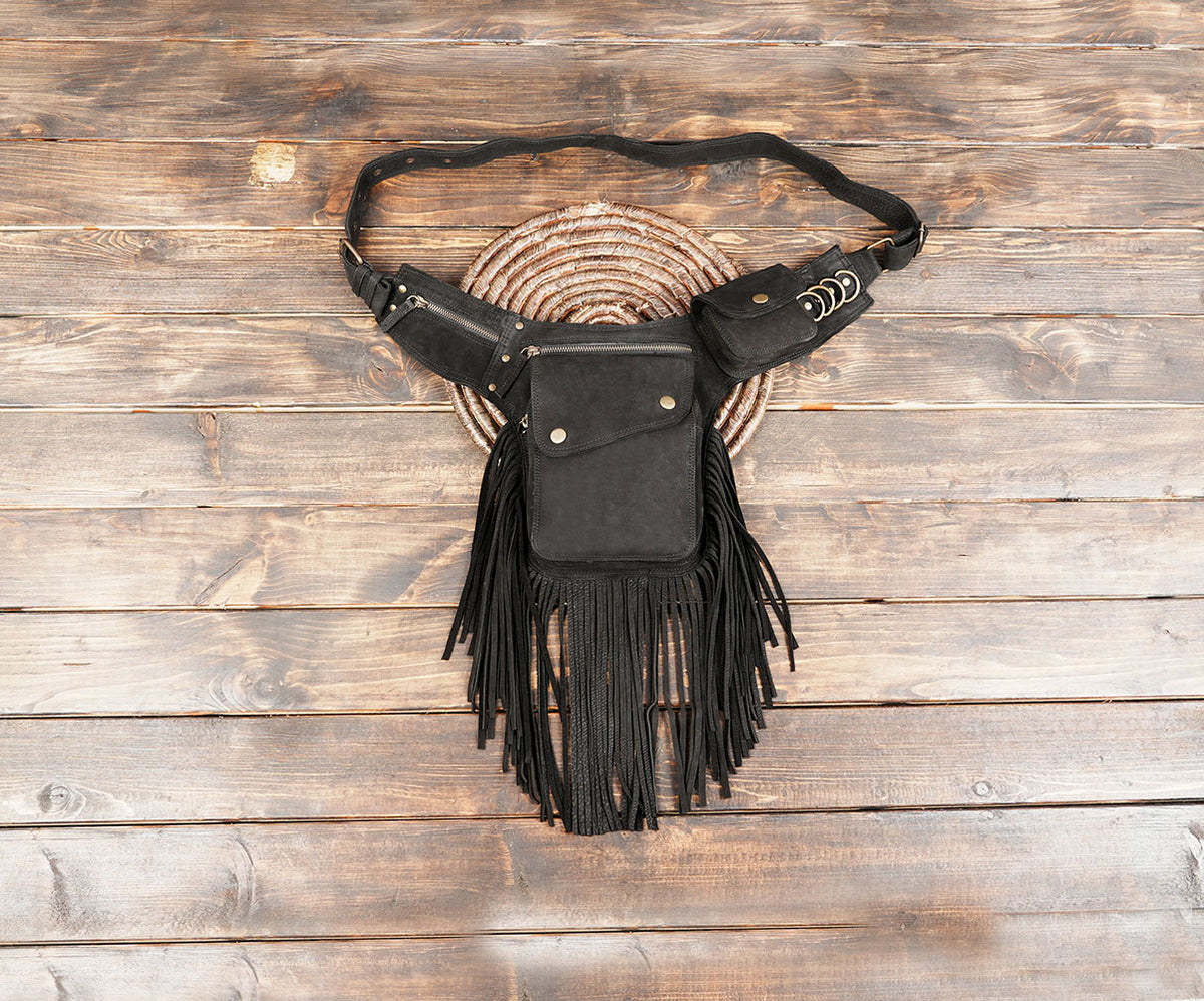 Black leather fringe waist bag with multiple pockets and brass-tone hardware on a rustic wooden background.