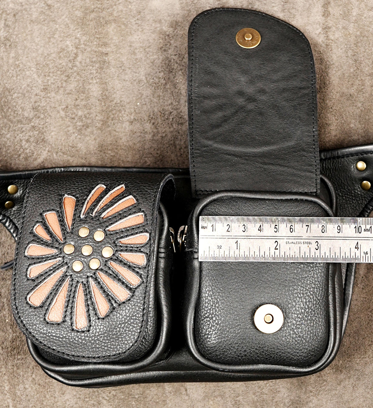 Black leather belt bag with a sunburst cutout pattern on the front pocket, shown open with a metal snap flap and a measuring tape laid across the front pocket to indicate size.