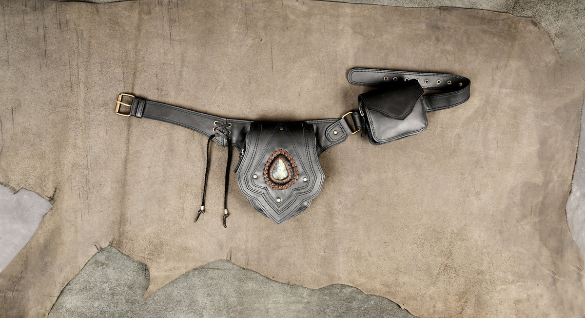 A black leather fanny pack with a central shield-shaped pouch featuring a decorative metallic emblem and dangling cords.