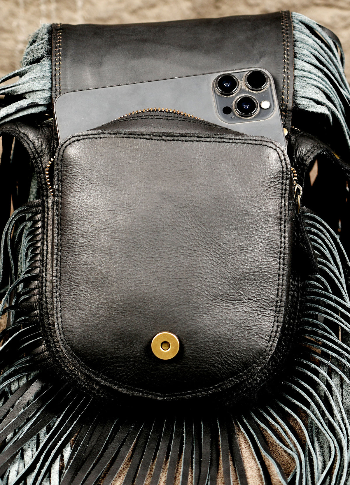 A close-up of a black leather crossbody bag with fringe detailing and a gold snap button, shown with a smartphone tucked into its side pocket.