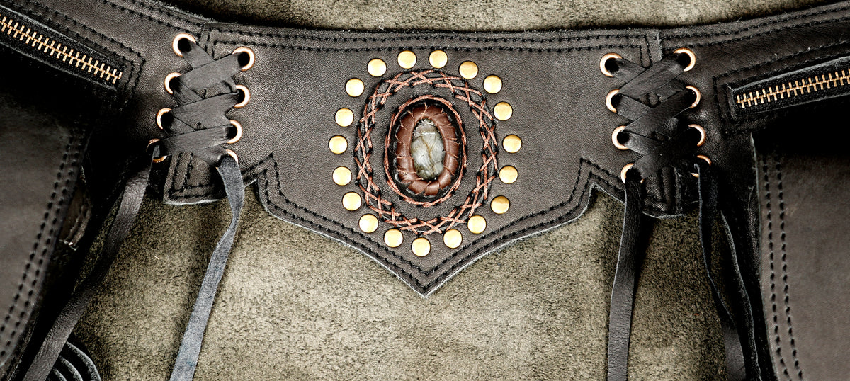 Close-up of a dark brown leather and suede vest with a central oval stone inlay, surrounded by brass studs and a braided leather border, with lace-up side details.
