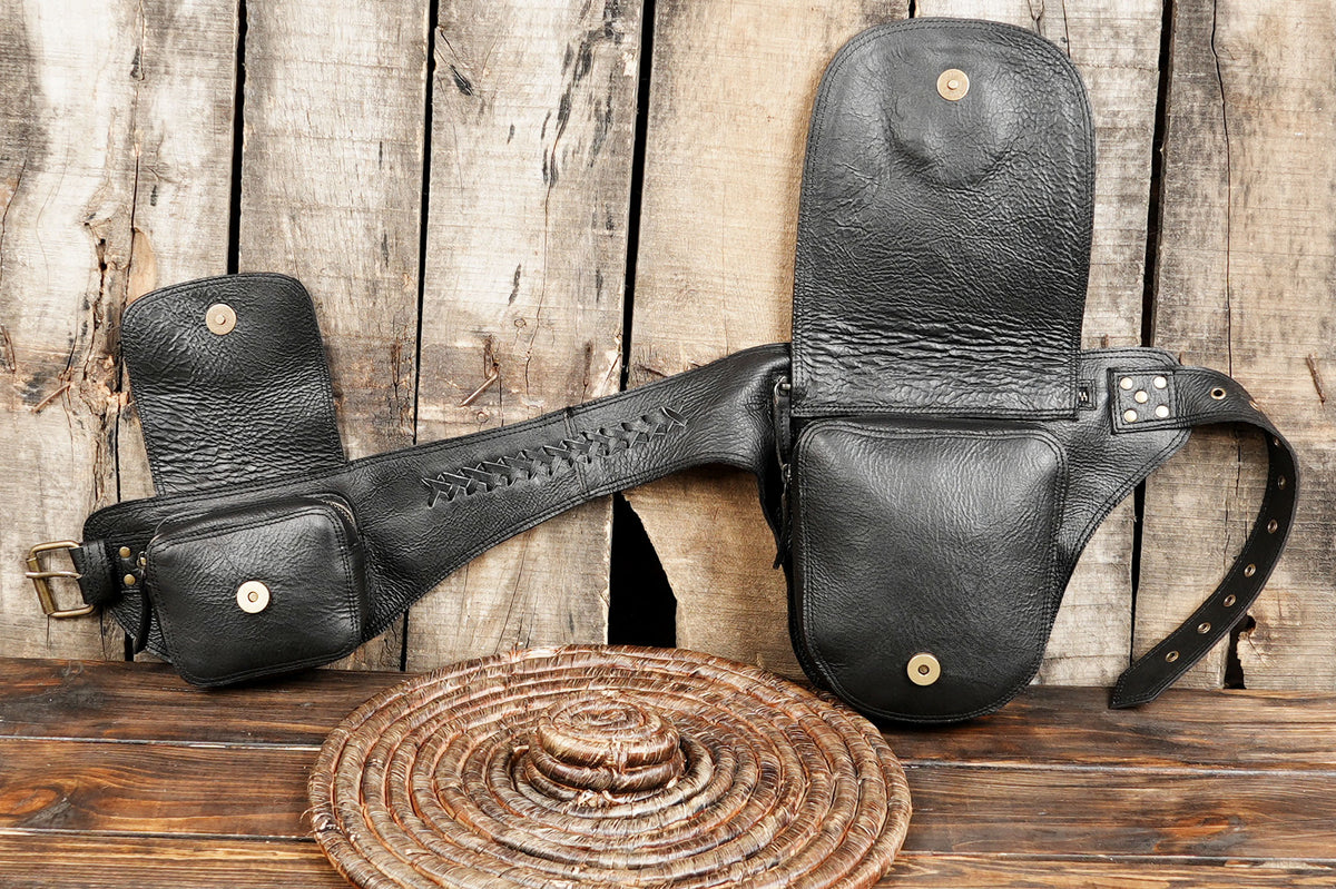 A black leather gun holster set with a matching belt and pouch, displayed on a rustic wooden surface with a woven hat in front.