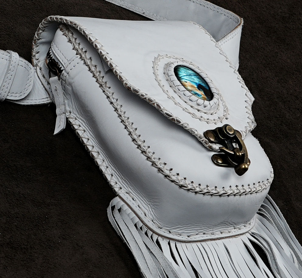 A white leather fringe pouch bag with a decorative blue and gold oval inlay on the flap, a brass clasp, and white braided stitching.