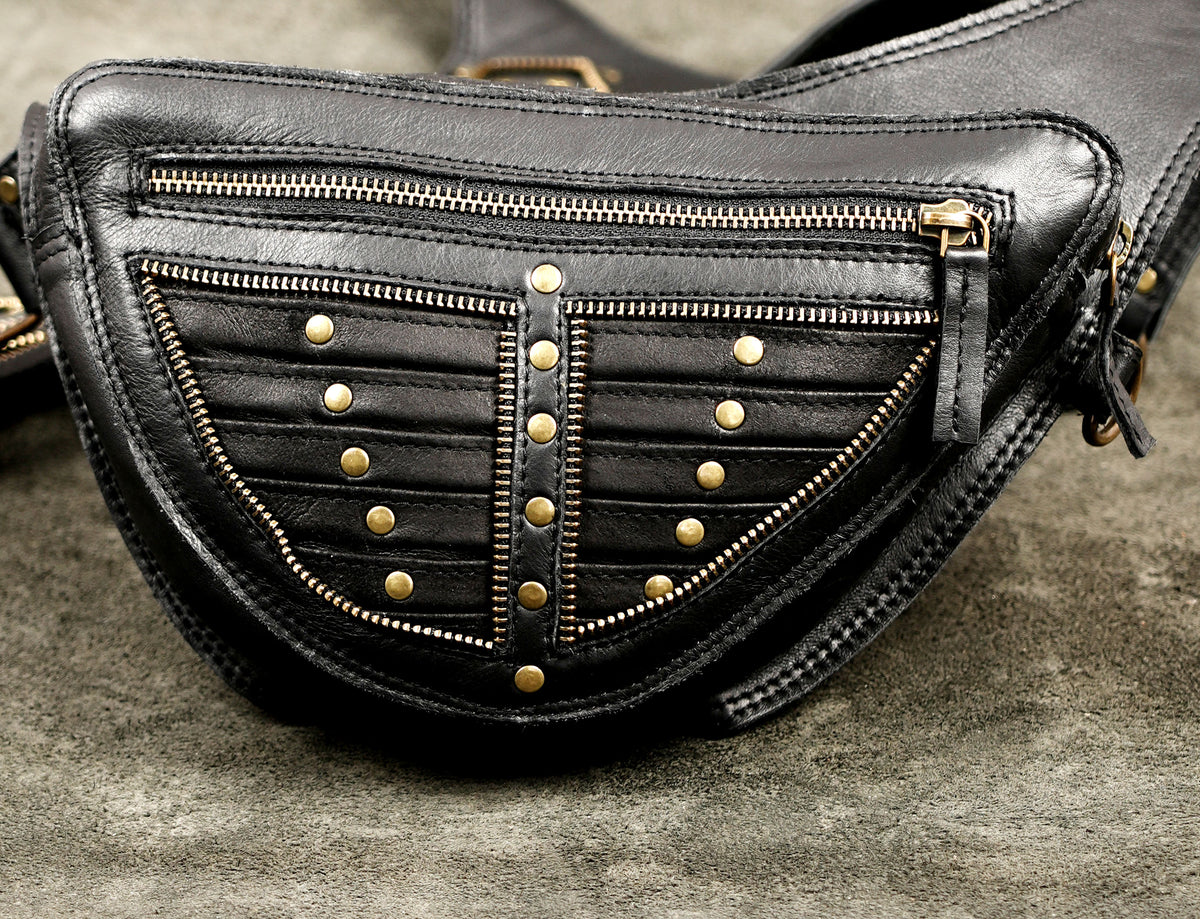 Black leather crossbody bag with gold-tone hardware, featuring multiple zippered compartments and decorative studs on a textured gray surface.