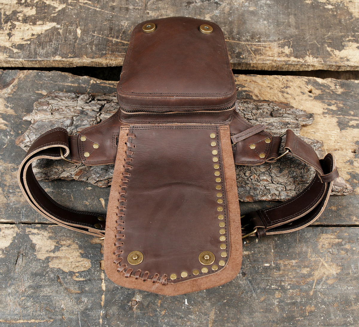 A brown leather crossbody bag with brass studs and a zippered top, displayed on a rustic weathered wooden surface.