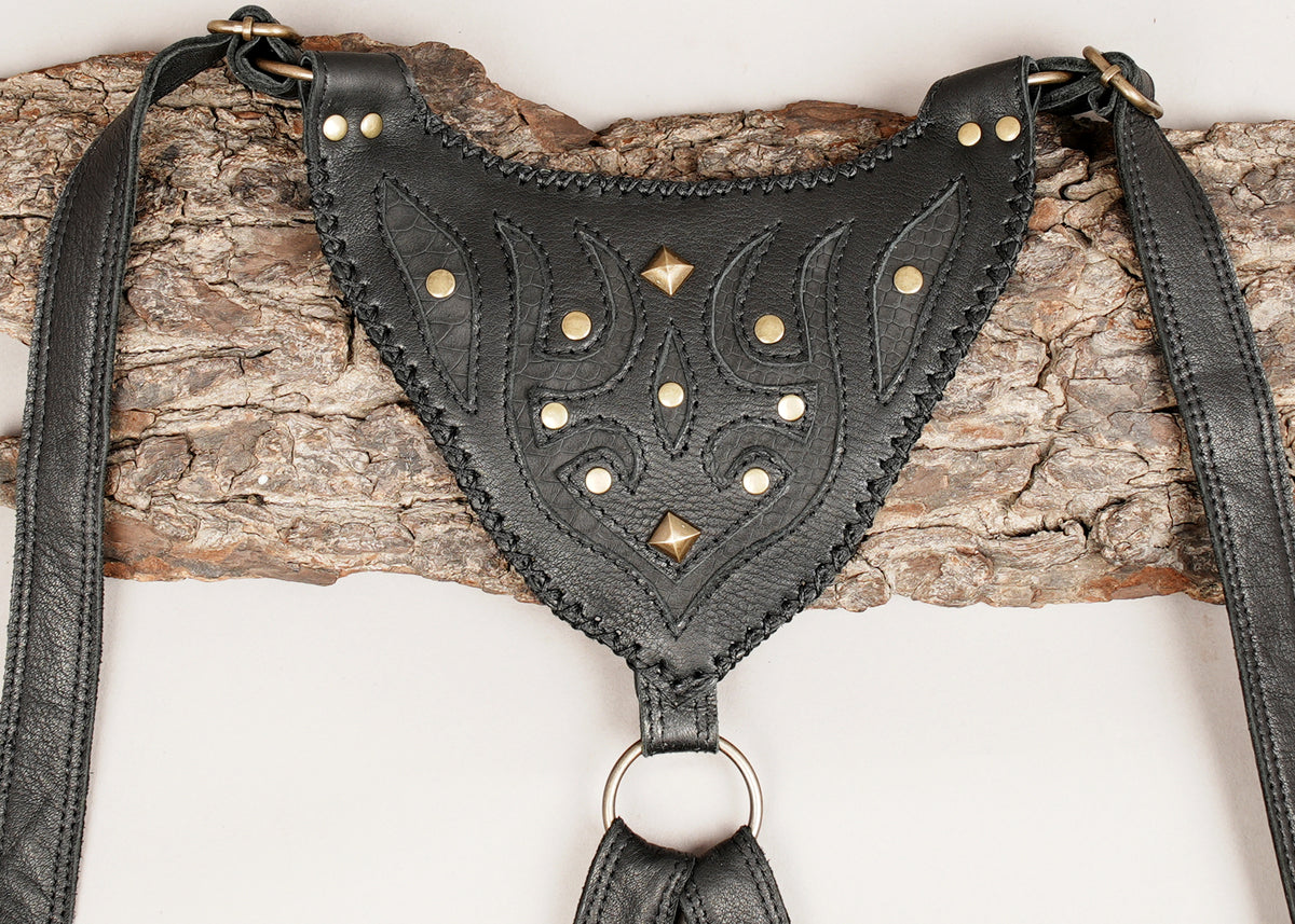 Black leather harness with decorative stitched patterns and brass studs, shown draped over a piece of tree bark.