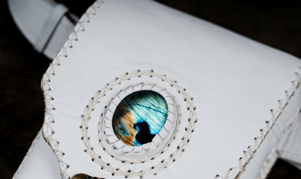 A close-up of a white leather accessory featuring a circular inlay with a blue, green, and brown iridescent pattern, surrounded by white braided stitching.
