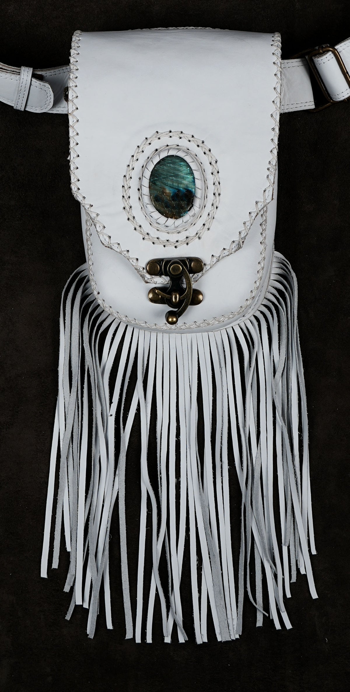White leather satchel bag with fringe, featuring a central oval abalone shell inlay and a brass buckle closure, shown against a dark background.
