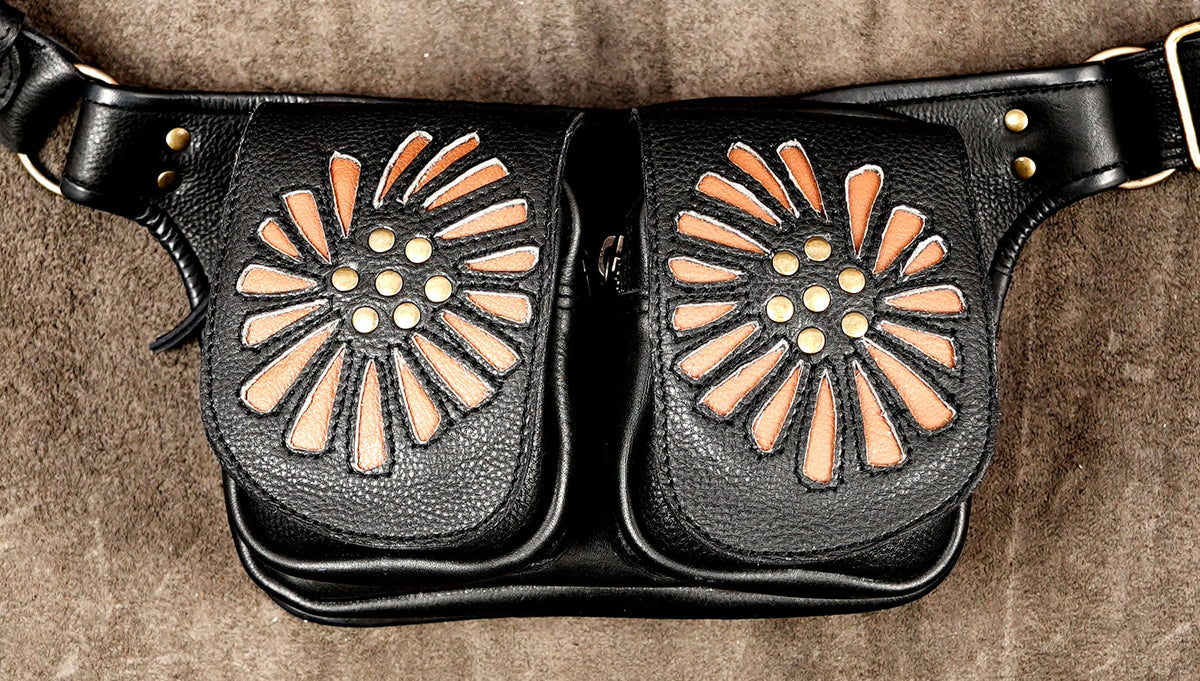 Black leather belt bag with two front pouches featuring sunburst cutouts filled with tan material and gold-tone studs, shown on a brown textured background.