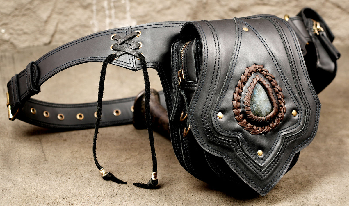 Black leather fanny pack with a wide adjustable strap, lace-up detail, and a decorative oval brooch featuring a blue-gray stone on the front flap.