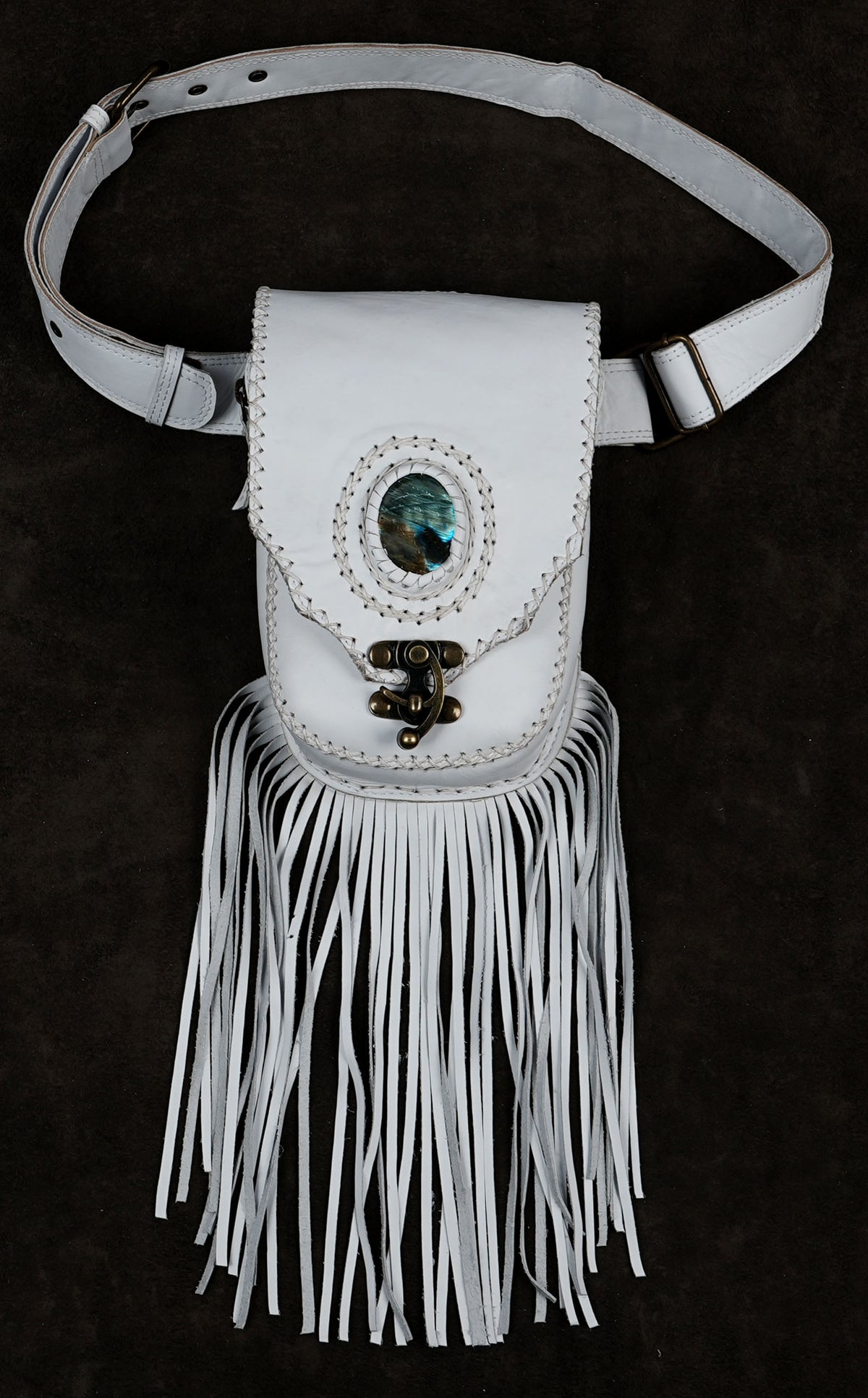 A white leather belt bag with a long fringe bottom, featuring a central oval turquoise stone and a decorative metal clasp, displayed against a dark textured background.