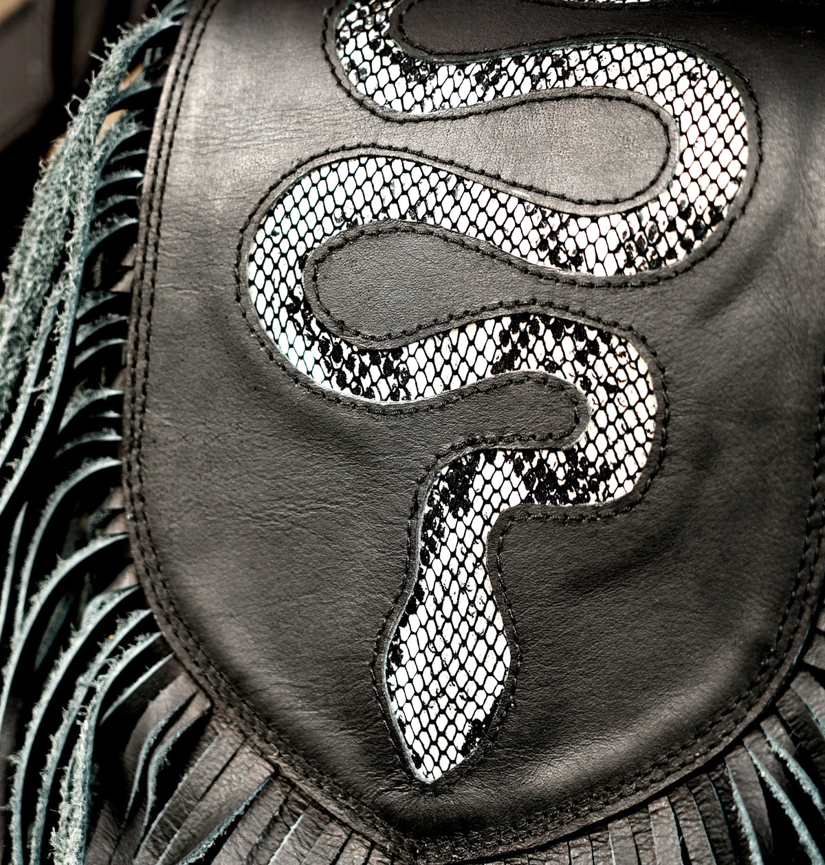 Close-up detail of a black leather bag featuring a large snake motif outlined in silver-toned mesh and surrounded by fringe.