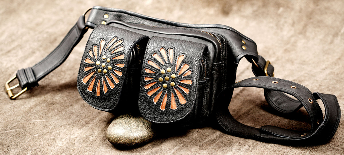 Black leather fanny pack with two front flap pockets featuring cutout floral patterns and brass studs, resting on a textured brown surface with a small stone.