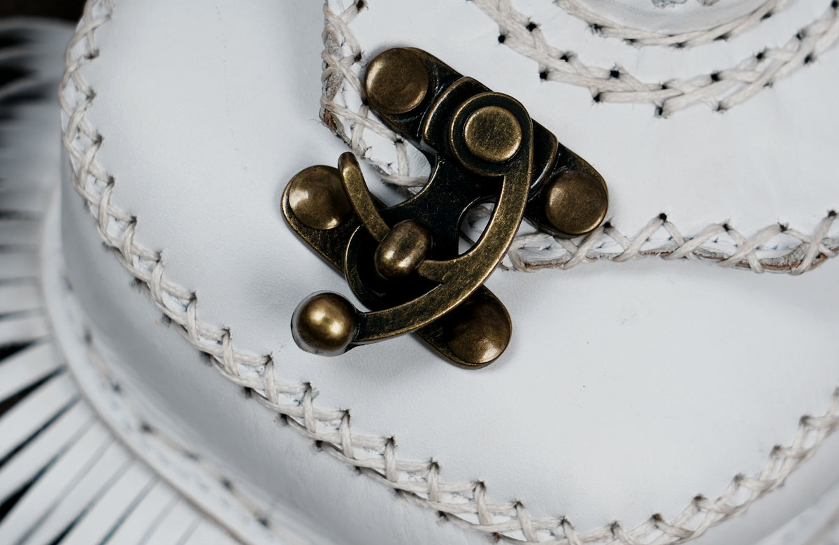 Close-up of a decorative white woven hat with a prominent antique bronze metal clasp on the front.