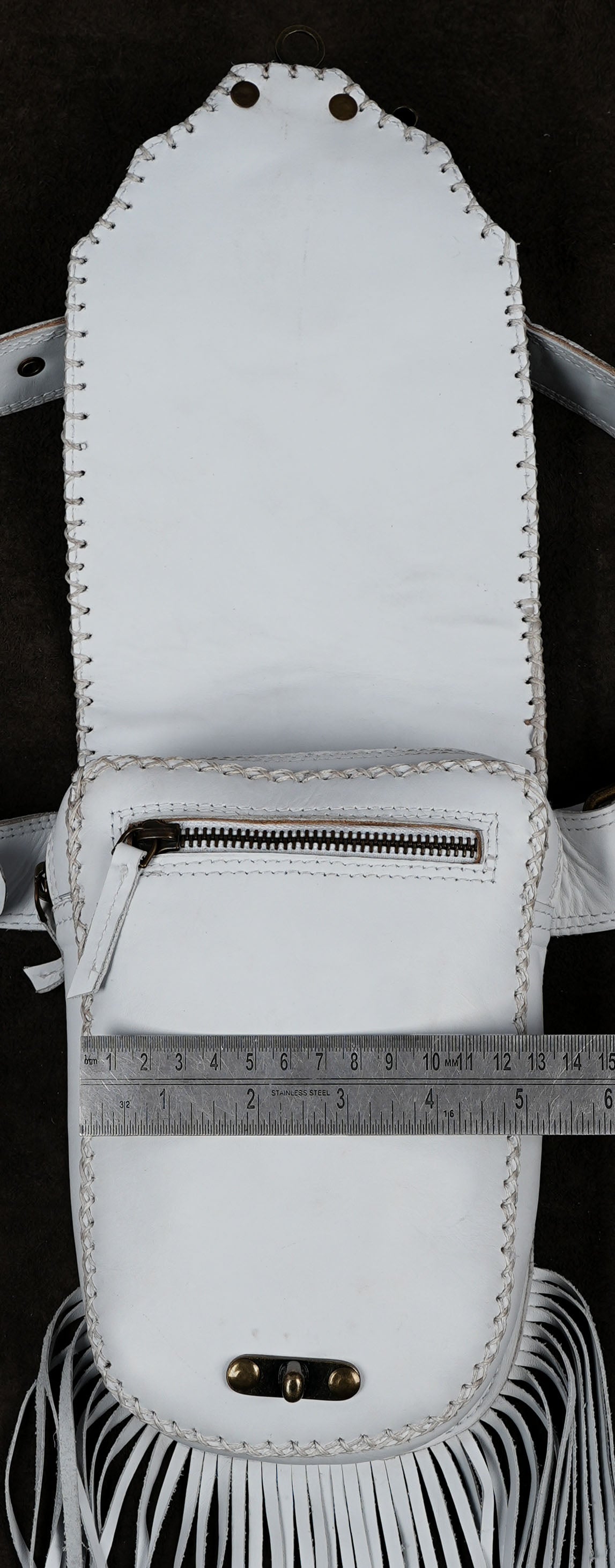 A white leather fringe pouch or small bag with a front zipper pocket, photographed against a dark background. A metal ruler is placed across the front to show the pouch's width.