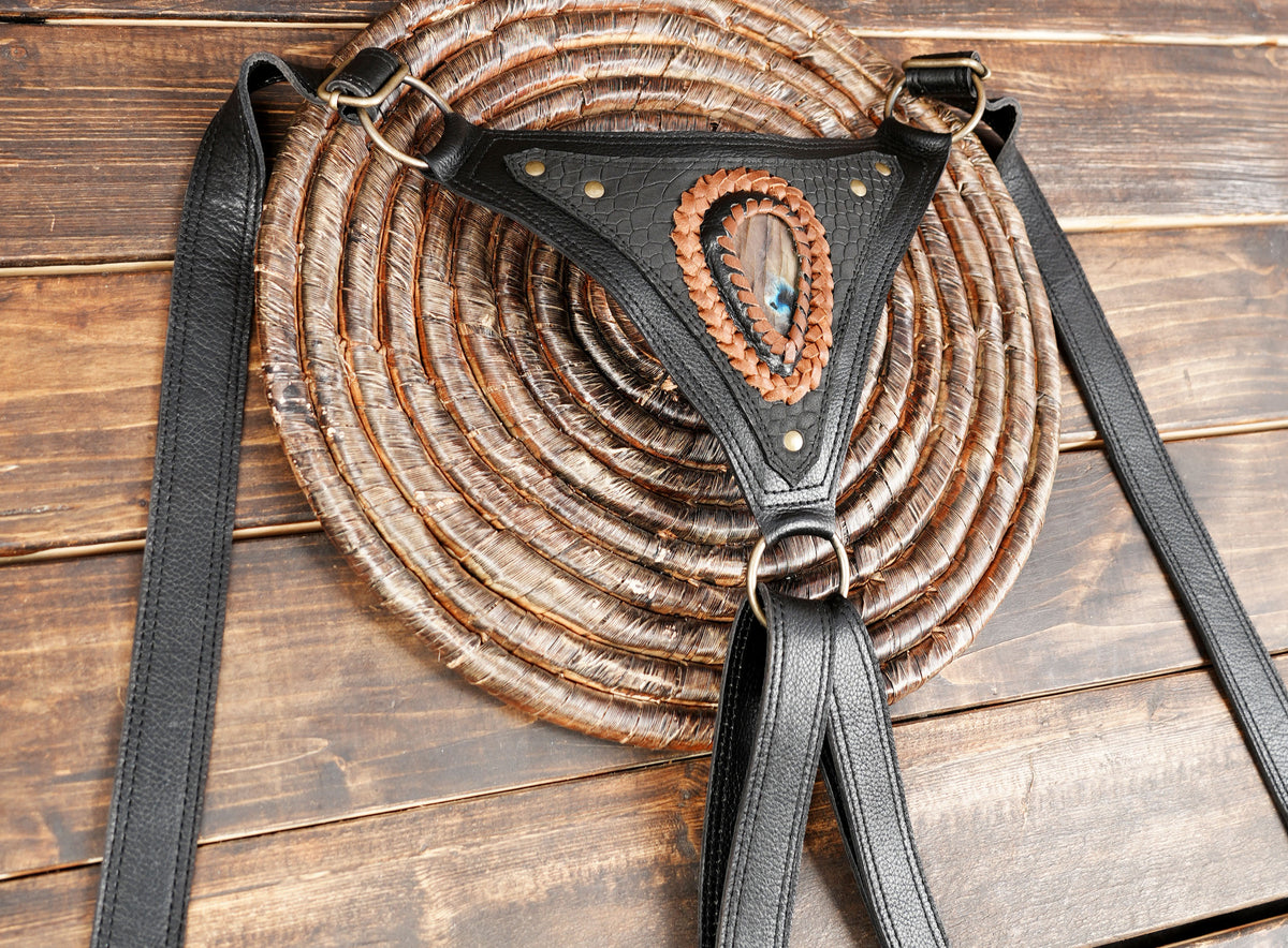 A black leather harness-style bag strap laid over a large round woven straw bag on a rustic wooden surface.