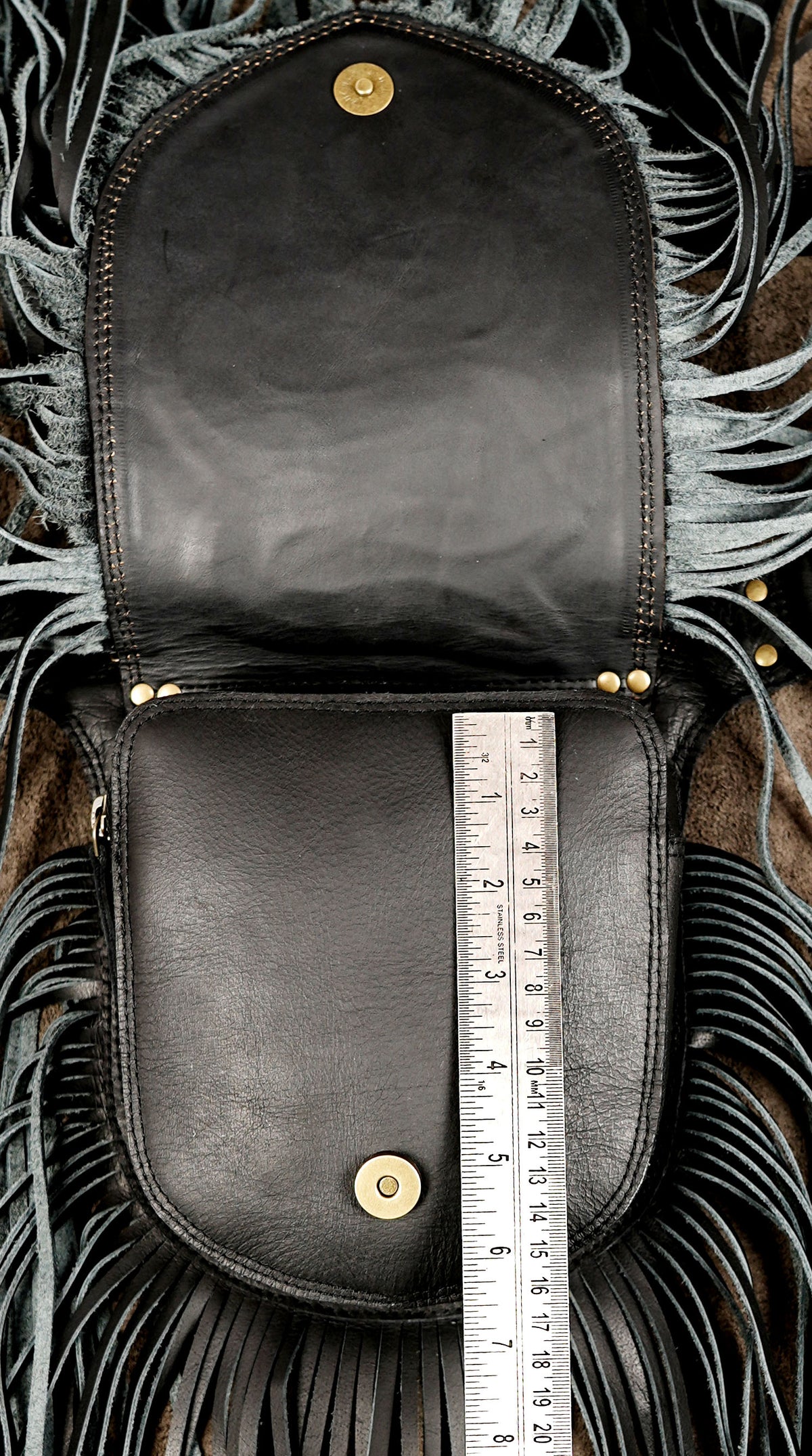 A black leather pouch or clutch shown open, with a metal ruler placed vertically inside the front pocket to indicate size. The bag has fringe detailing around the edges and gold-tone snap hardware.