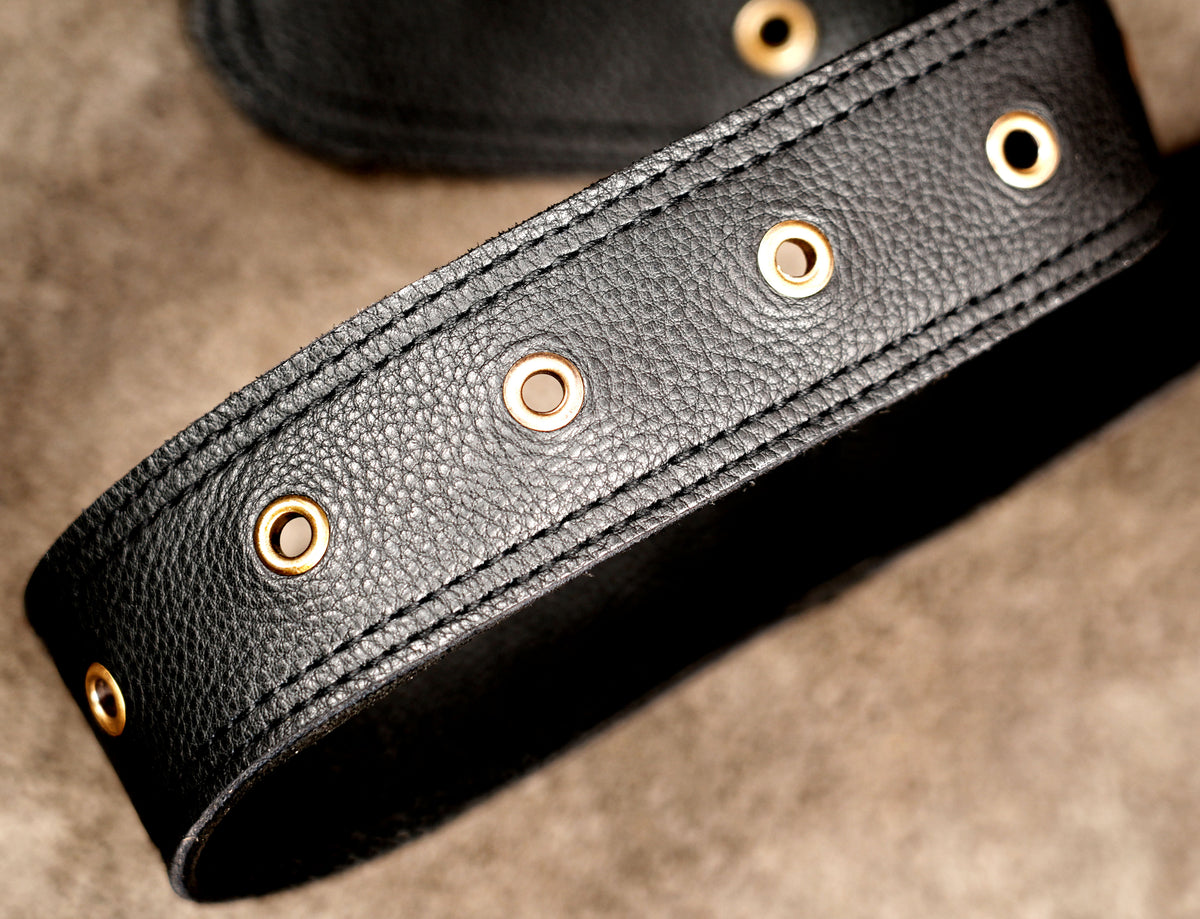 A close-up of a black textured leather belt with gold-tone metal grommets, resting on a neutral brown textured surface.