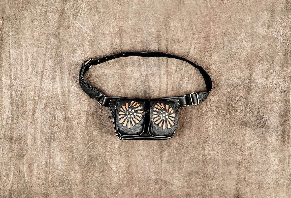 A black leather fanny pack with two front pockets decorated with circular sunburst patterns in tan and orange, resting on a textured brown surface.