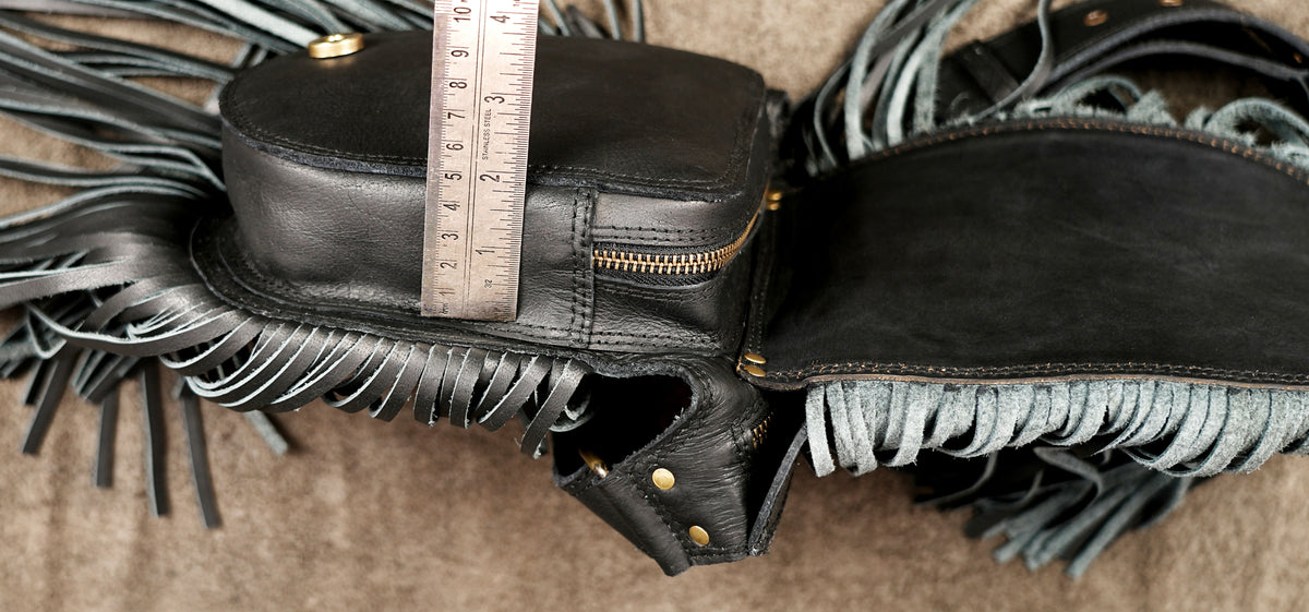 A black leather fringe bag shown open on a textured tan surface, with a metal ruler placed vertically inside the main compartment to indicate size.