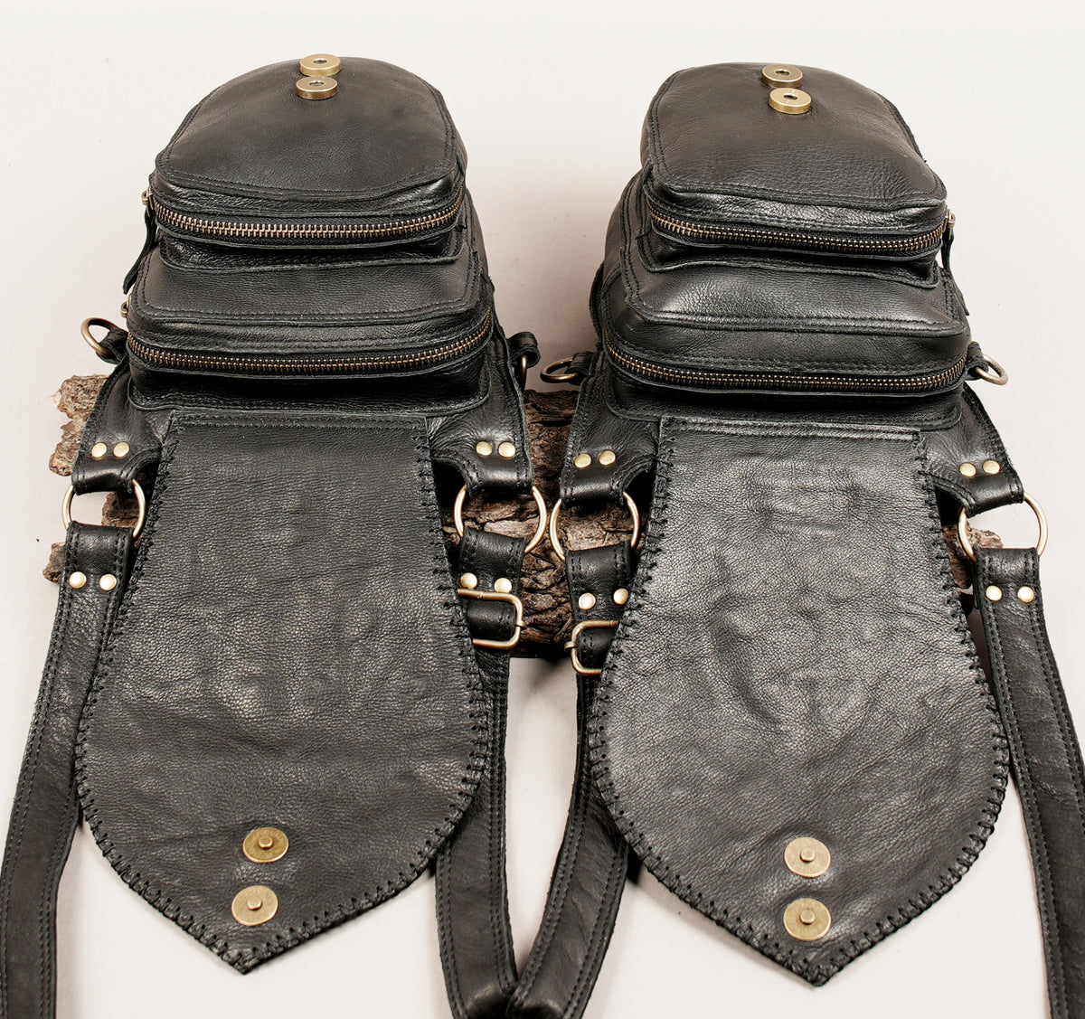 A pair of black leather crossbody bags with multiple zippered compartments, brass-tone hardware, and pointed flap details.