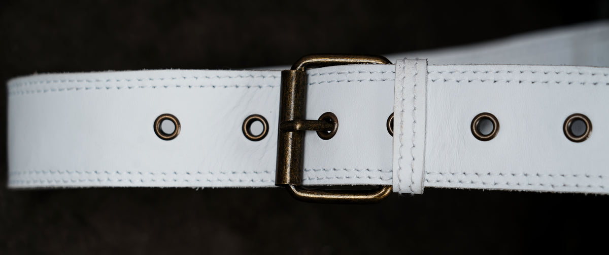 A close-up of a white leather belt with a brass buckle and metal eyelets, shown against a dark background.
