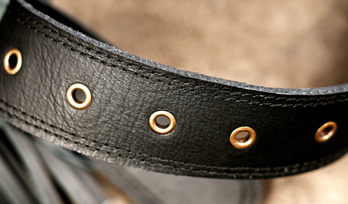 A close-up of a black leather belt with brass-tone metal grommets, showing the textured leather grain and stitching.