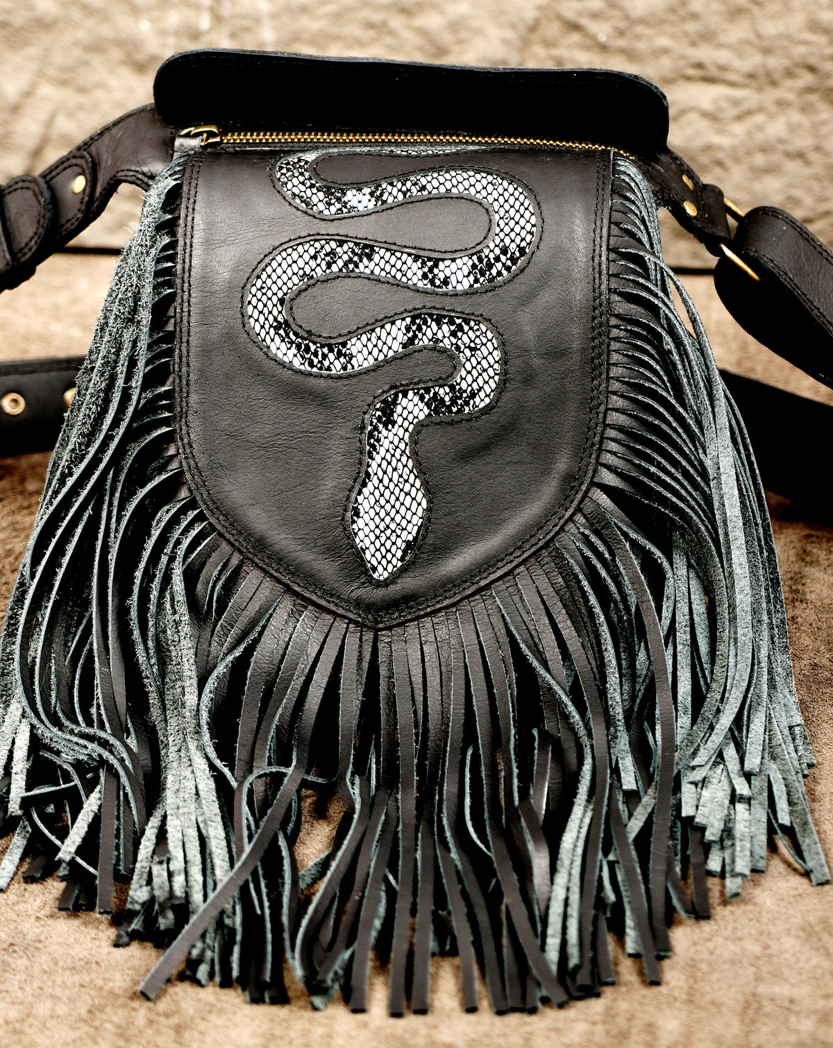 Black leather fringe crossbody bag featuring a shield-shaped front pocket with a raised snake motif in black and white snakeskin print.
