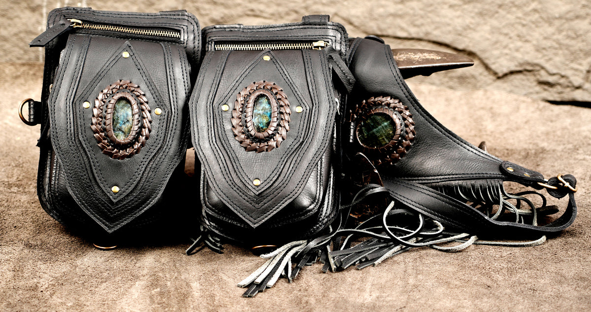 Black leather dual holster pouch bag displayed flat on textured backdrop with brass hardware and fringe.