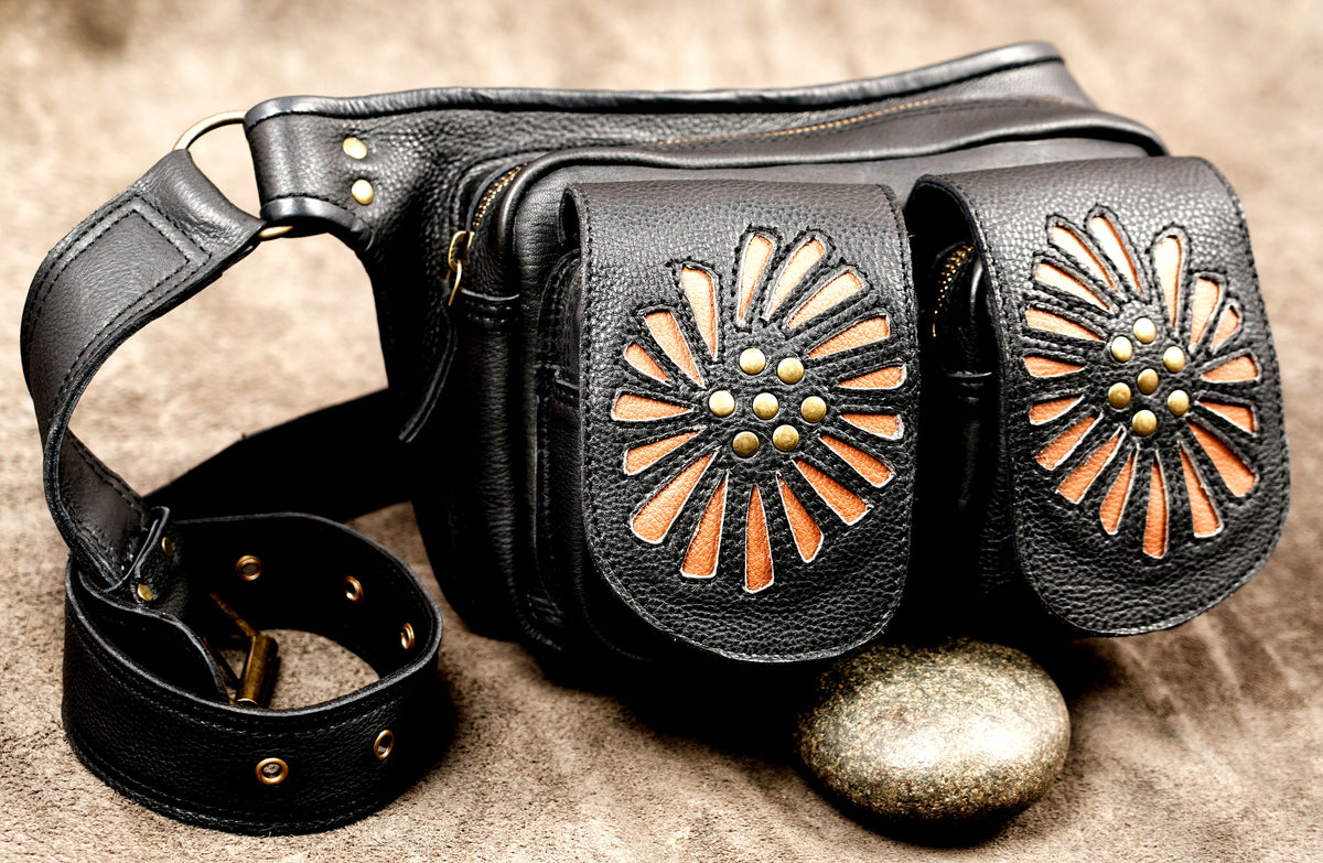 Black leather fanny pack with two front flap pockets featuring cutout sunburst designs in tan and brass studs, shown on a textured brown surface with a stone prop.