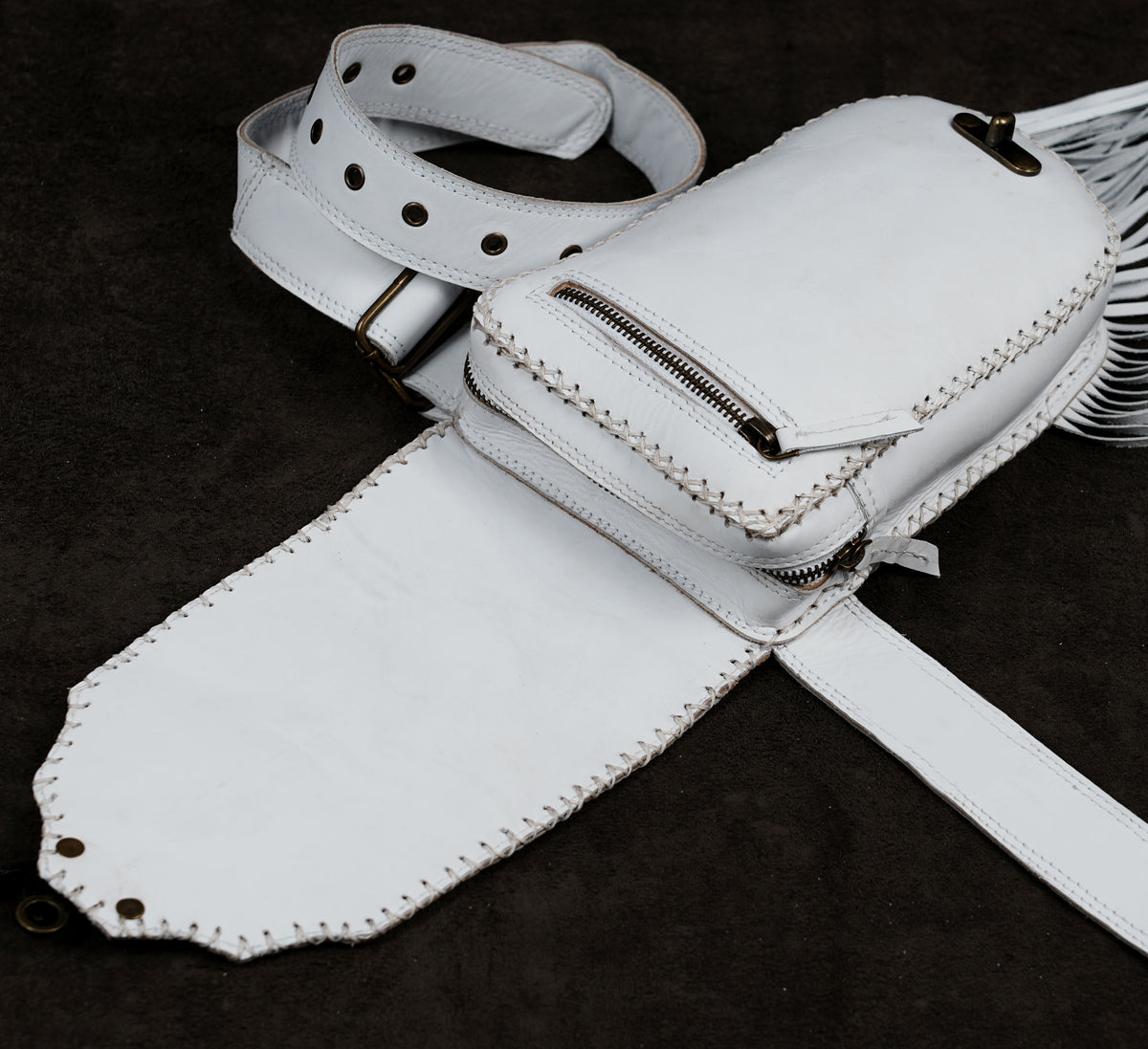 A white leather belt bag with fringe detailing, shown laid flat on a dark textured background.