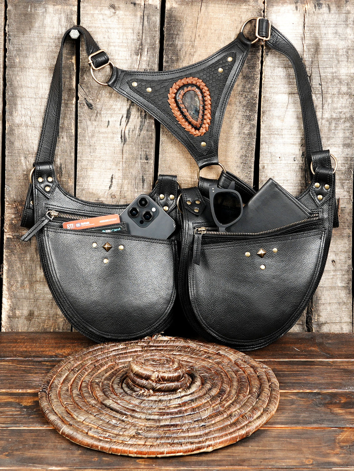 A black leather shoulder bag with a triangular harness-style strap and multiple front pockets, shown on a rustic wood plank surface with a woven mat in front. The bag has brass-tone hardware, a circular woven accent on the strap, and accessories placed in the pockets, including a phone, card, glasses, and wallet.