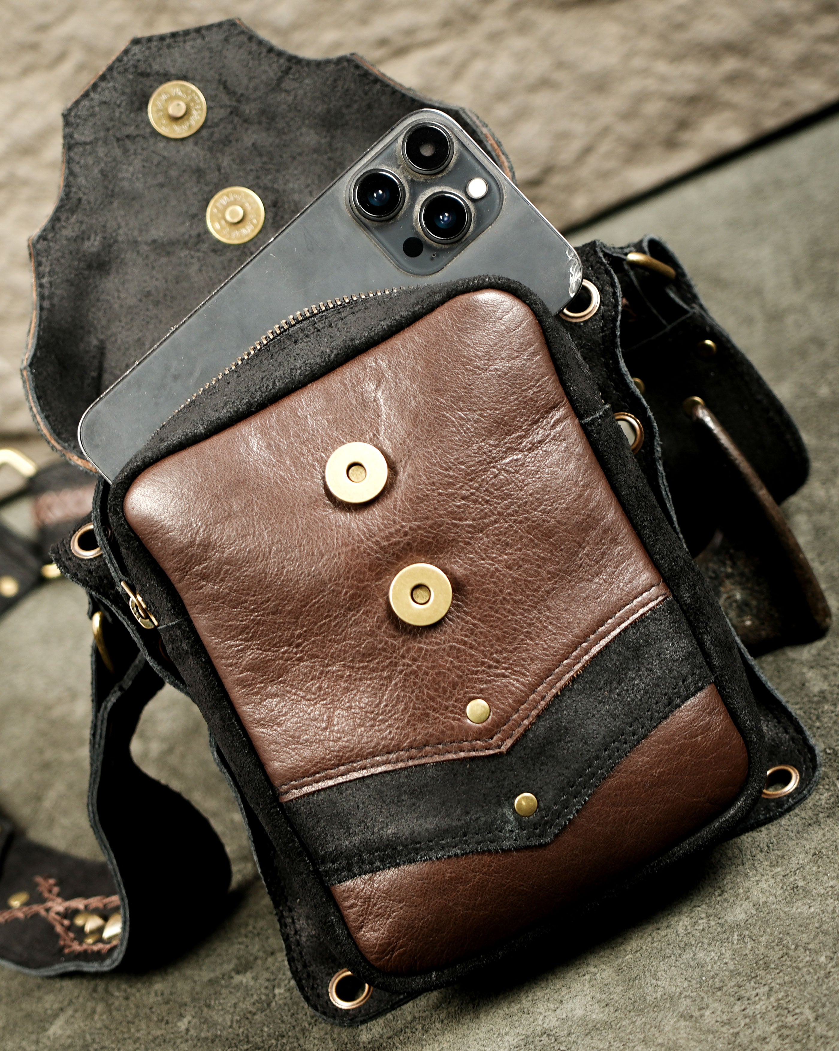 A brown and black leather crossbody bag with brass-tone snap buttons and hardware, shown open with a smartphone tucked inside, on a textured gray surface.