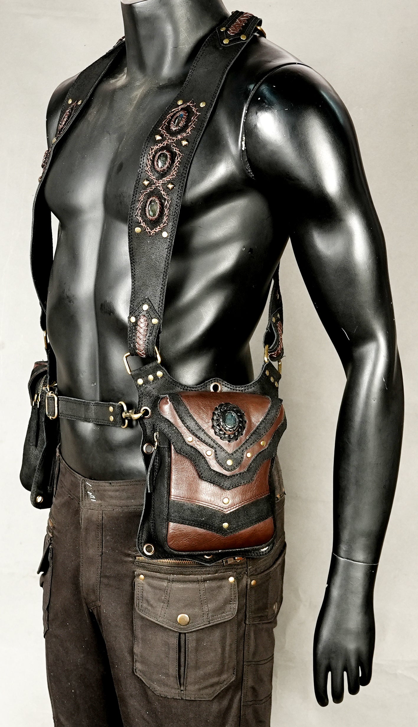 A steampunk-style brown leather crossbody pouch with black straps and ornate metal hardware, displayed on a black mannequin torso wearing dark cargo pants against a light gray background.