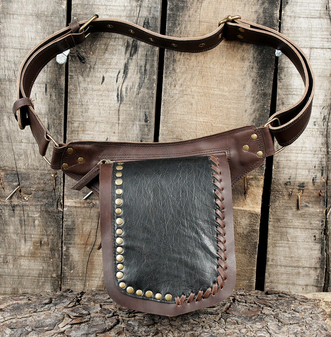 A brown leather belt bag with a large front pouch, studded trim, and a wide adjustable belt, displayed against a rustic wooden plank background.