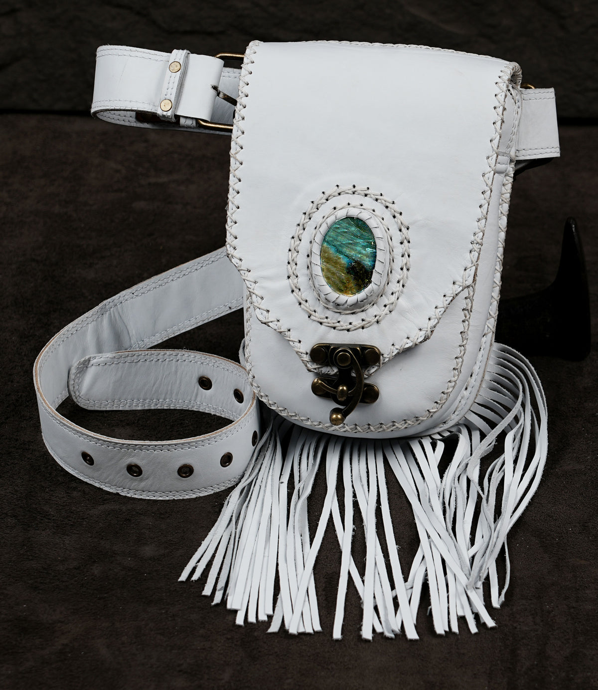 A white leather fringe crossbody bag with a decorative oval green stone and brass-tone hardware.