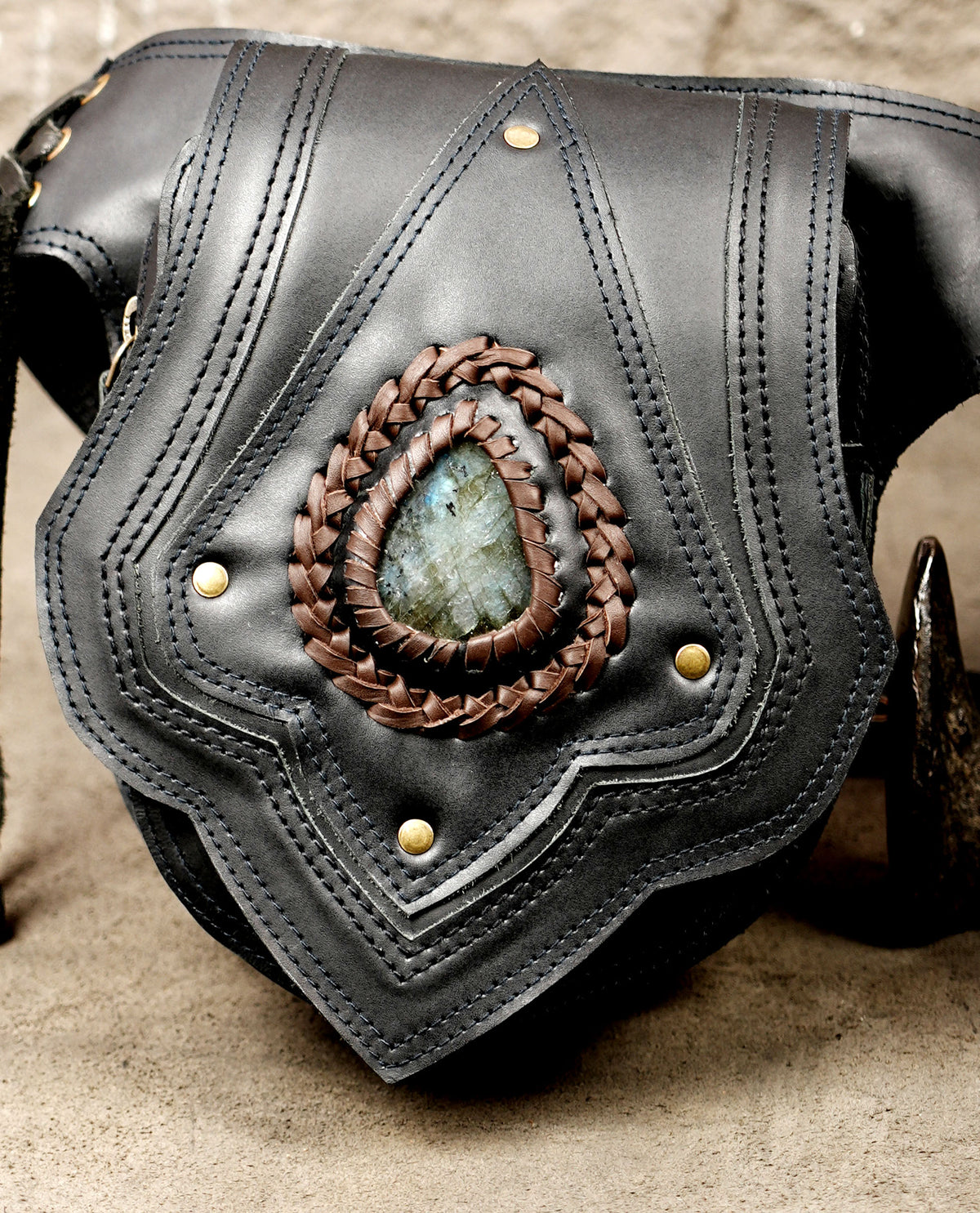 A close-up of a black leather shoulder bag featuring a central oval inlay with a braided brown leather border and a blue-gray stone. The bag has brass-tone rivets.