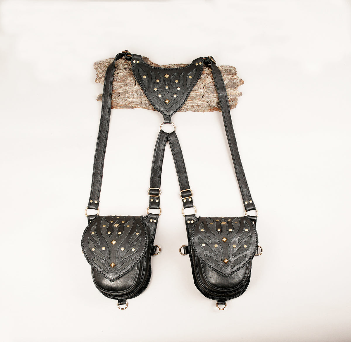 Black leather suspender-style crossbody bags with studded detailing, displayed on a light wood branch against a plain white background.