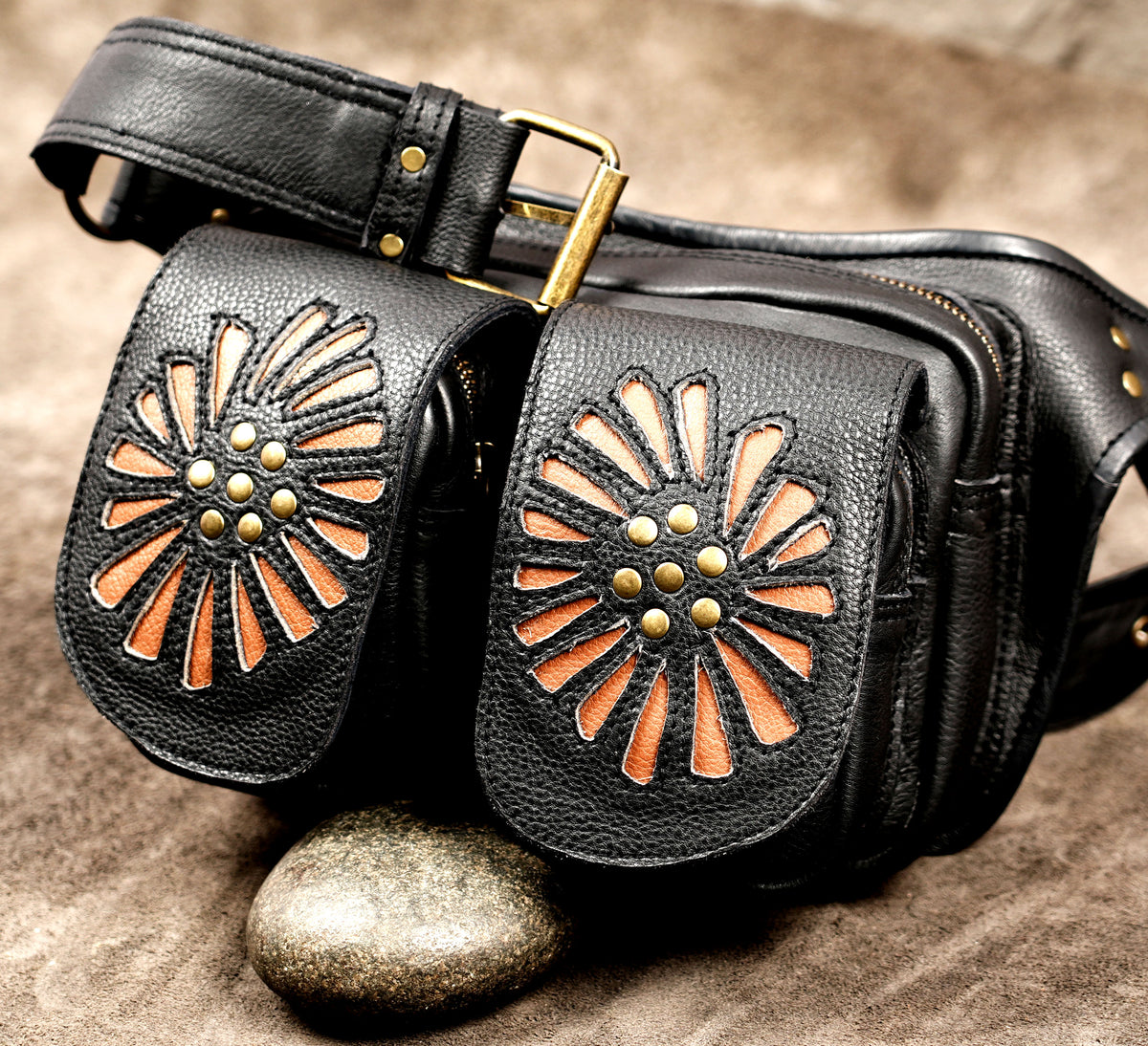 Black leather fanny pack with two front pouches featuring cutout sunburst designs in orange-brown and gold-tone studs, photographed on a textured brown surface with a stone prop.
