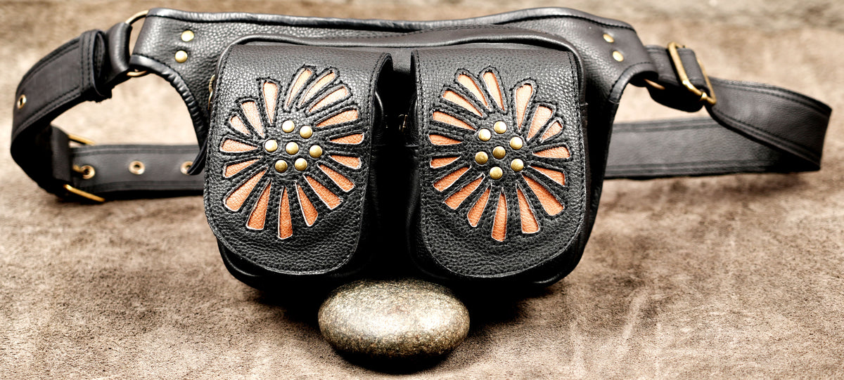 Black leather belt bag with two front pouches decorated with tan and brown sunburst patterns and gold-tone studs, displayed on a textured brown surface with a stone in front.