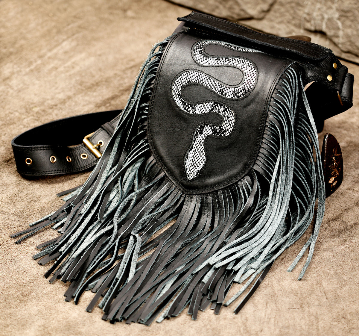 Black leather fringe pouch bag with a silver snake graphic on the front flap, resting on a textured tan surface.