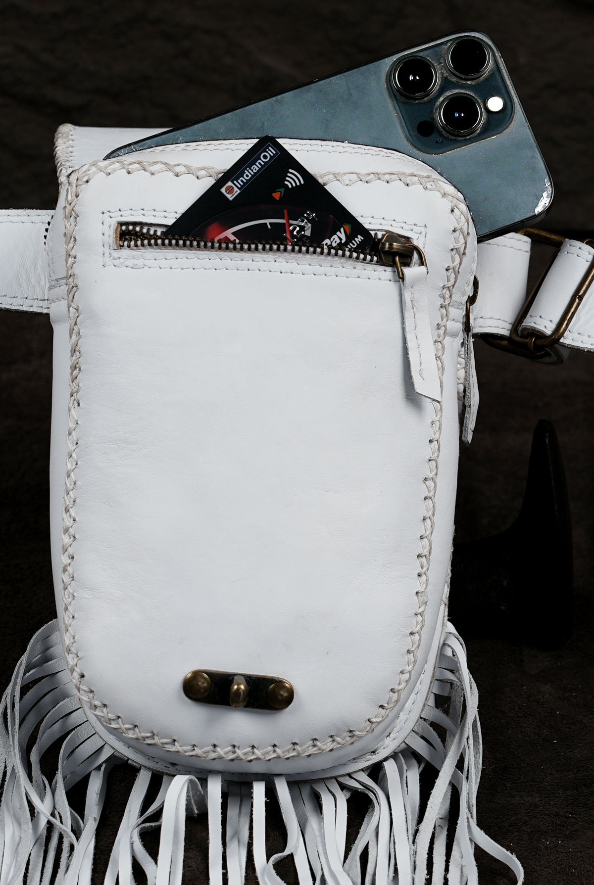 A white leather crossbody bag with fringe detailing and a front zip pocket, shown with a smartphone tucked into the top pocket against a dark textured background.