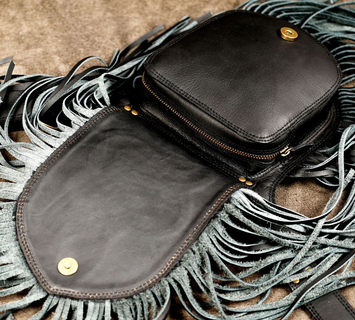 A black leather crossbody bag with a large flap and long fringe, laid flat on a textured brown surface with matching fringe around it.