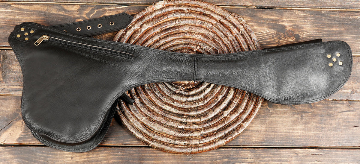 A black leather saddlebag-style case with a zipper closure and metal studs, laid flat on a rustic wooden surface over a round woven mat.