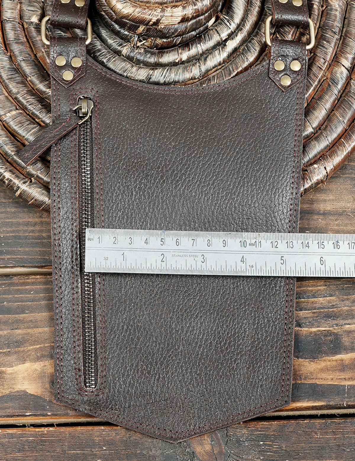 Dark brown textured leather pouch with a vertical zipper, shown with a metal ruler measuring its width on a rustic wooden background.