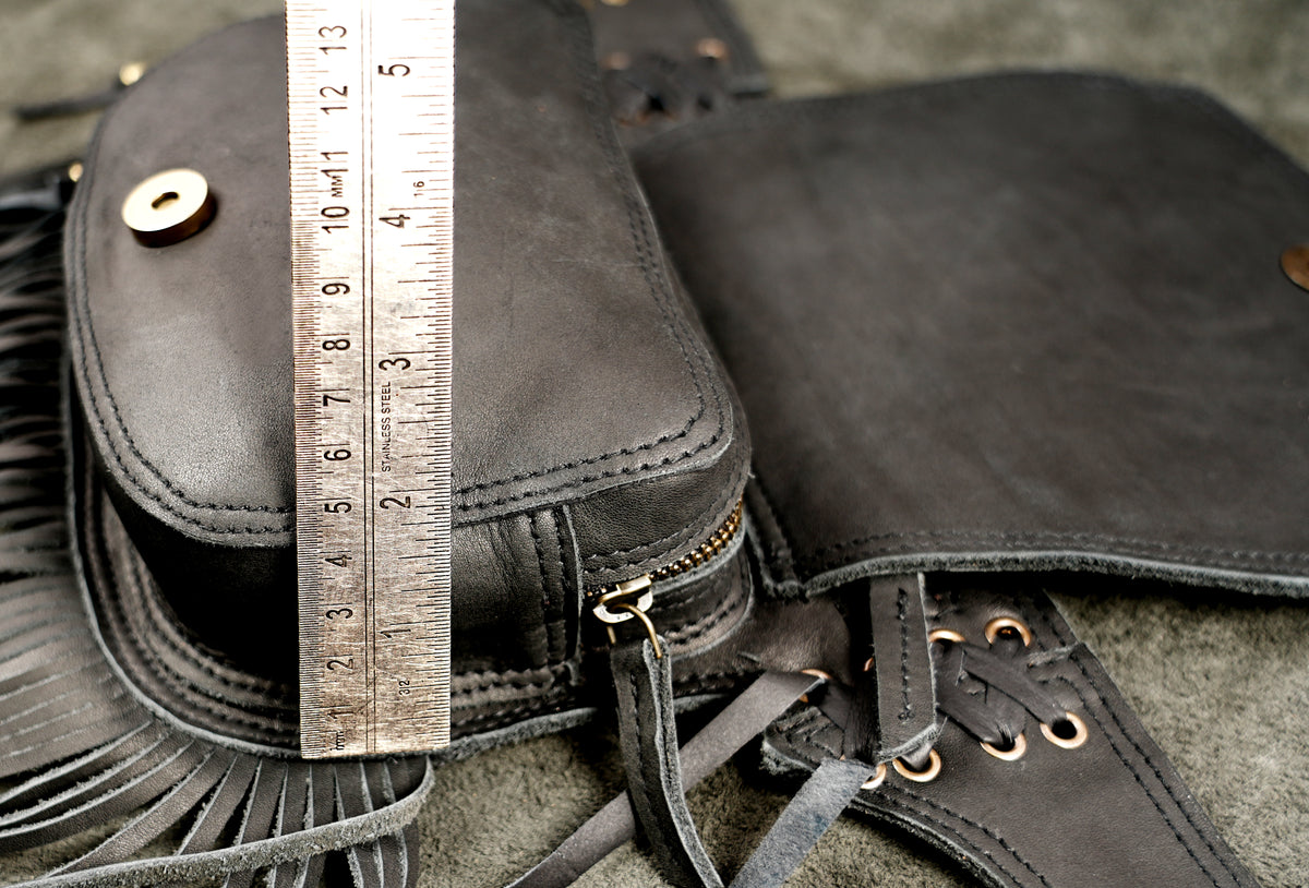 Close-up of a black leather bag with fringe, a zipper, lace-up strap, and a metal ruler placed vertically to show dimensions.