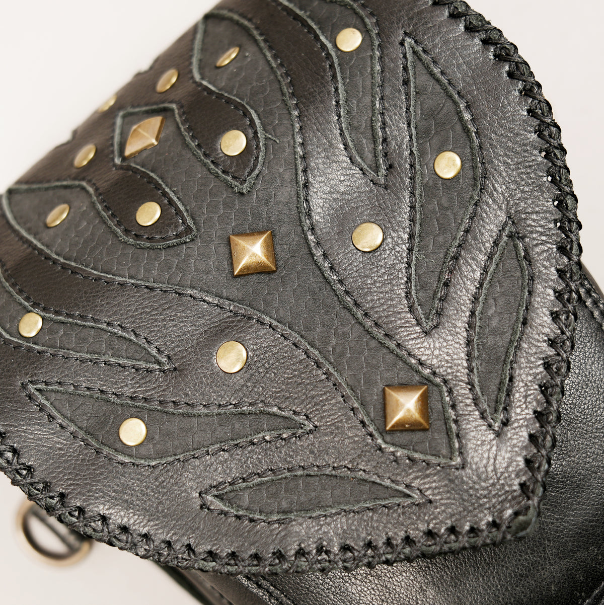 Close-up of a dark gray textured leather accessory featuring stitched leaf-like cutouts, gold-tone studs, and pyramid studs on a light background.
