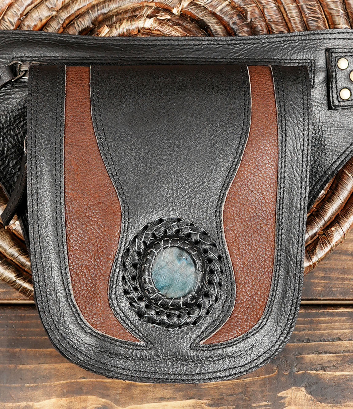 A close-up of a black leather pouch with brown leather side panels and a central circular woven detail featuring a blue-gray stone, shown on a rustic wooden surface with a woven mat in the background.