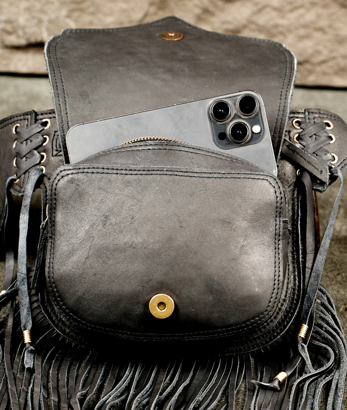 A dark distressed leather shoulder bag with fringe and lace-up details, shown holding a gray smartphone in its front slip pocket .