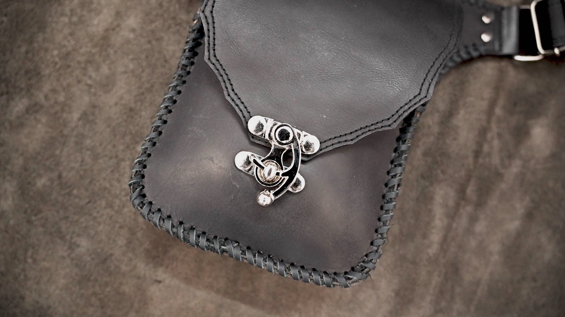 Handmade black leather thigh bag with adjustable waist strap on a textured background.